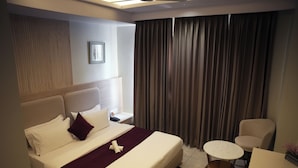 A hotel room with a large bed, a small round table, a chair, and a window with curtains. - Tr Hotels (Ambala)