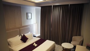 A hotel room with a large bed, a small round table, a chair, and a window with curtains. - Tr Hotels (Ambala)