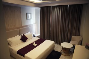 A hotel room with a large bed, a small round table, a chair, and a window with curtains. - Tr Hotels (Ambala)