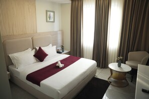 A hotel room with a large bed, a bedside table with a vase, a chair, and a small round table. - Tr Hotels (Ambala)