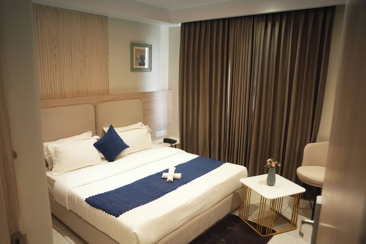 A hotel room with a bed, a small table with a vase, and a chair.