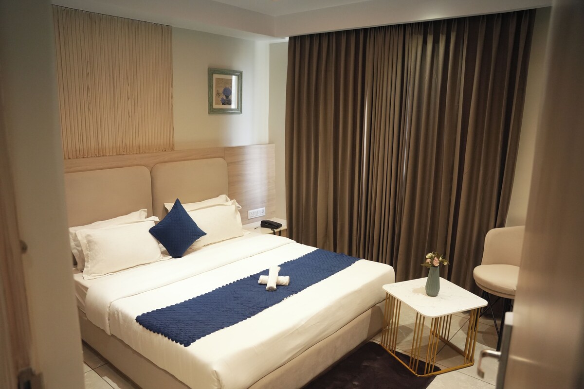 A hotel room with a bed, a small table with a vase, and a chair.