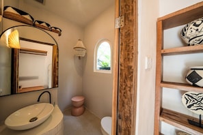 Ein Badezimmer mit einem weißen Waschbecken, einem runden Spiegel, einer Pendelleuchte, einem kleinen Fenster und einem Holzregal mit Dekoration.