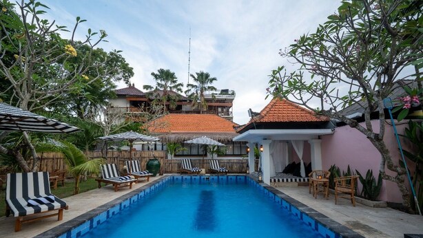 A swimming pool with lounge chairs and umbrellas, surrounded by tropical plants and buildings with red-tiled roofs. - Casa Coco (Lembongan Island)