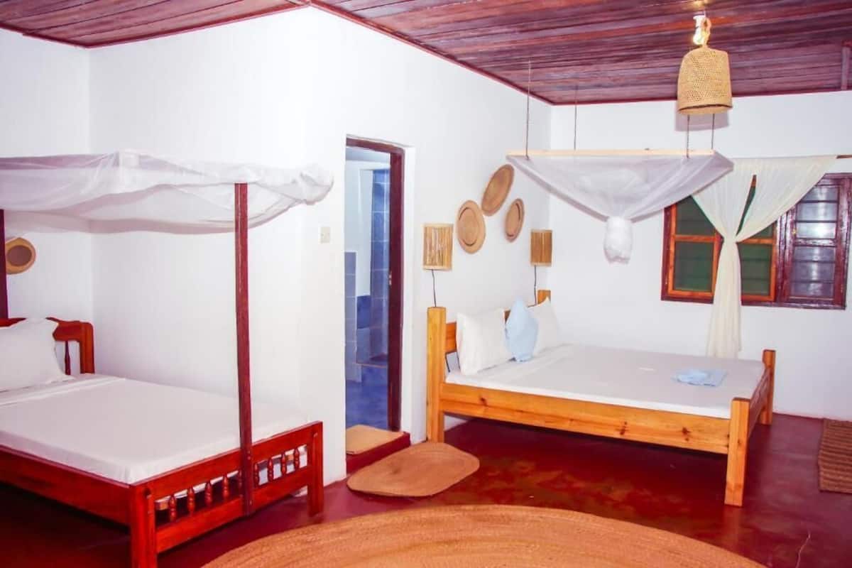 Una habitación con dos camas de madera, una con dosel y otra sin. Hay una cesta tejida colgando del techo, un taburete pequeño de madera y un felpudo en el suelo.