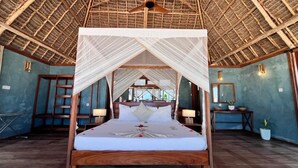 A four-poster bed with a canopy, wooden headboard, bedside tables with lamps, and a view through the thatched roof to the outside. - Tiki Beach Club & Resort (Paje)