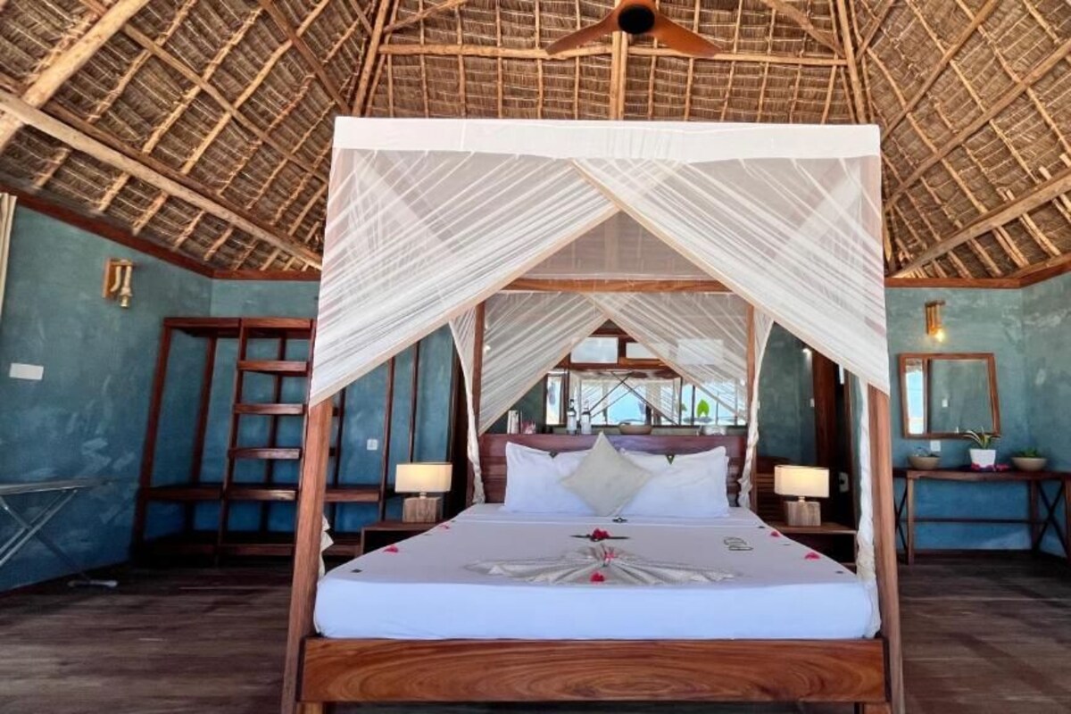 A four-poster bed with a canopy, wooden headboard, bedside tables with lamps, and a view through the thatched roof to the outside.