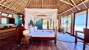 A room with a bed, a small table, a nightstand, a window, and a view of the ocean. - Tiki Beach Club & Resort (Paje)