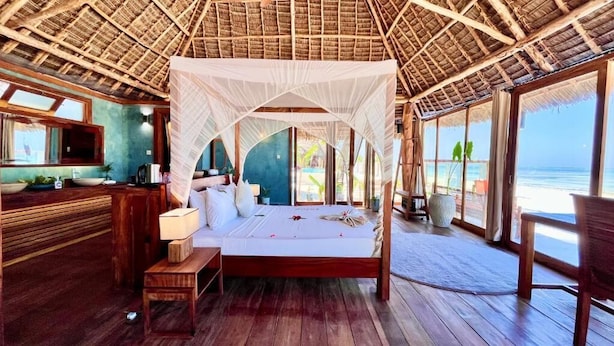 A room with a bed, a small table, a nightstand, a window, and a view of the ocean. - Tiki Beach Club & Resort (Paje)