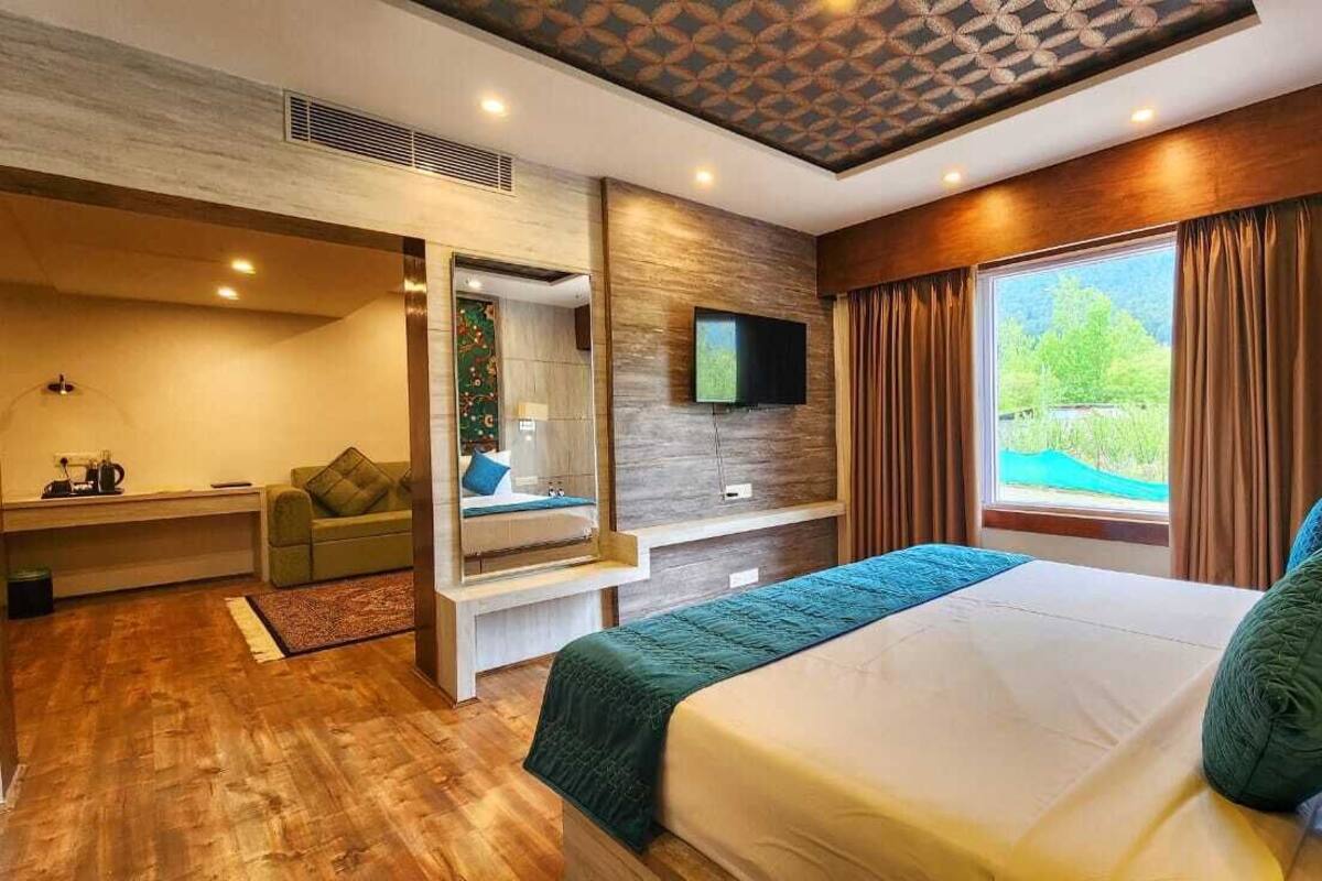 A modern hotel room with a large bed, a TV mounted on the wall, a seating area with a sofa and a coffee table, and a view of a pool through the window.