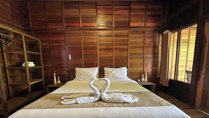 Una cama doble con ropa de cama blanca y dos toallas con forma de cisne, paneles de madera en las paredes, una mesita de noche con velas y una ventana con cortinas.