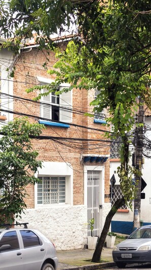 Um edifício residencial com paredes de tijolo, uma porta branca e uma janela com grade de metal. Carros estão estacionados na frente e uma árvore com folhas verdes no primeiro plano.
