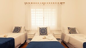 Une chambre d’hôtel avec un grand lit, deux petits lits, une fenêtre avec des rideaux légers et une table de chevet avec une lampe.