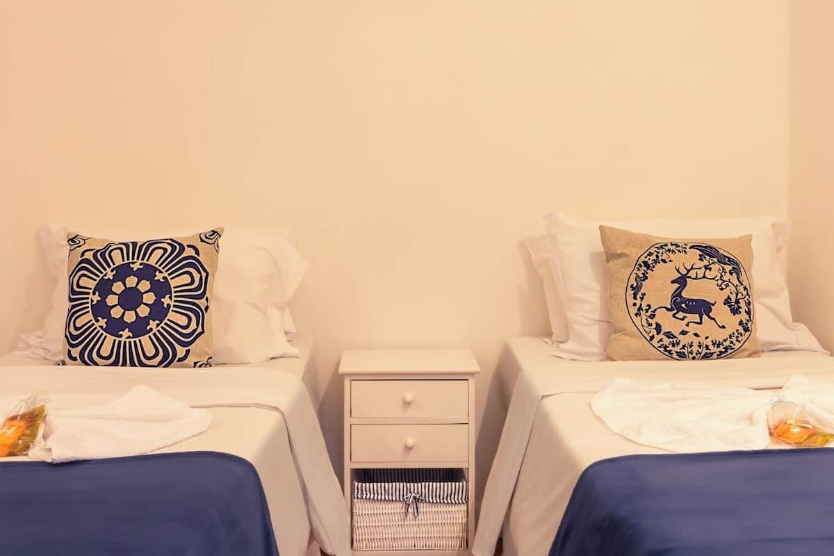 A hotel room with two beds, each with a white bedspread and a decorative pillow. There is a small bedside table with a drawer and a wicker basket.
