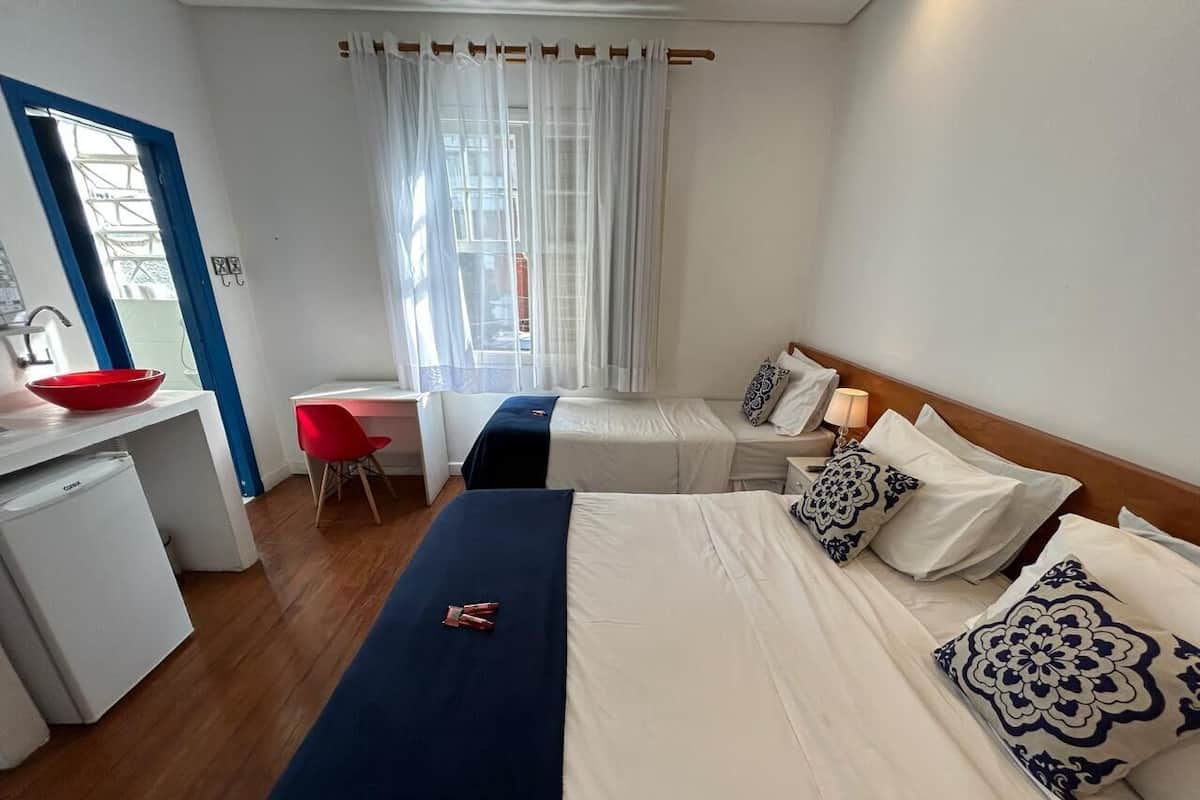 A hotel room with a bed, a desk, a red chair, a small refrigerator, and a window with curtains.
