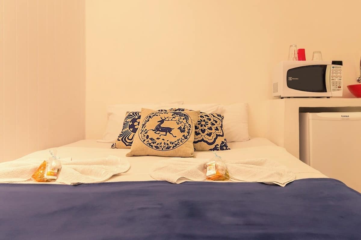 A neatly made bed with a blue bedspread, two decorative pillows, a small white bedside table with a red bowl and a microwave, and a white refrigerator.