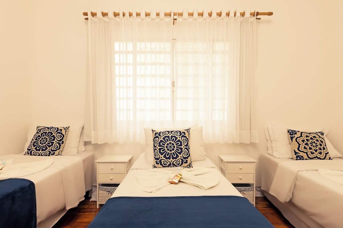 A hotel room with two single beds, a large window with sheer curtains, and a bedside table with a lamp.