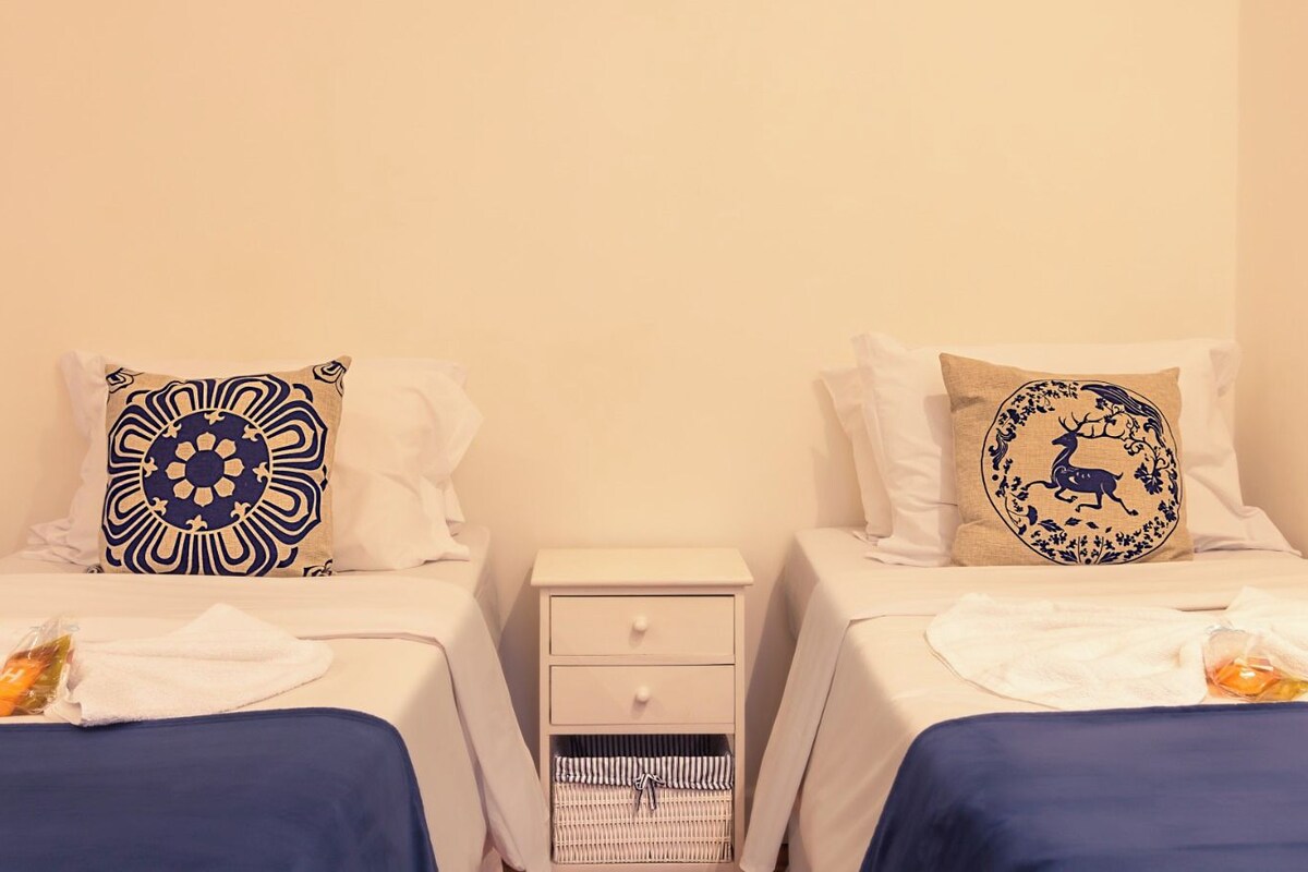 Een hotelkamer met twee bedden, elk voorzien van een witte spreidek en een decoratief kussen. Er is een klein nachtkastje met een lade en een rieten mandje.