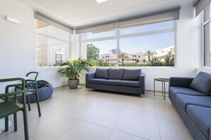 A modern living room with a grey sofa, a small round table, and a green chair. There is a large window with a view of buildings and trees. - Dunas Hostel & Guesthouse (Portimão)