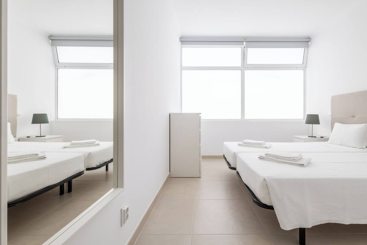 A hotel room with two beds, a large window, a bedside table with a lamp, and a wardrobe.