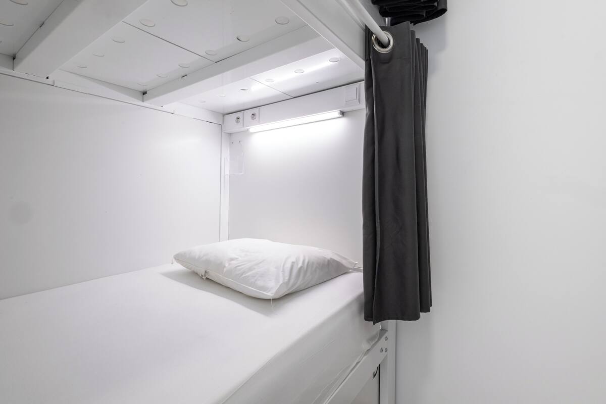 A single bed with a white pillow and a black curtain in a small, white room with a ceiling light.