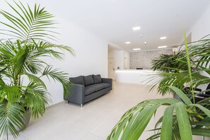 A modern living room with a grey sofa, a kitchen area in the background, and large green plants.