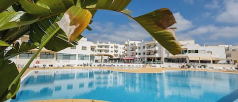 A swimming pool with a clear blue water surface, surrounded by a pebble-lined edge. In the background, there are white buildings with multiple balconies and windows.