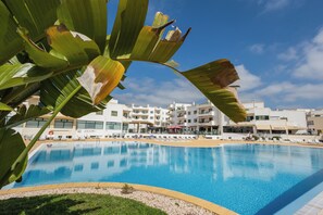 A swimming pool with a clear blue water surface, surrounded by a pebble-lined edge. In the background, there are white buildings with multiple balconies and windows. - Dunas Hostel & Guesthouse (Portimão)