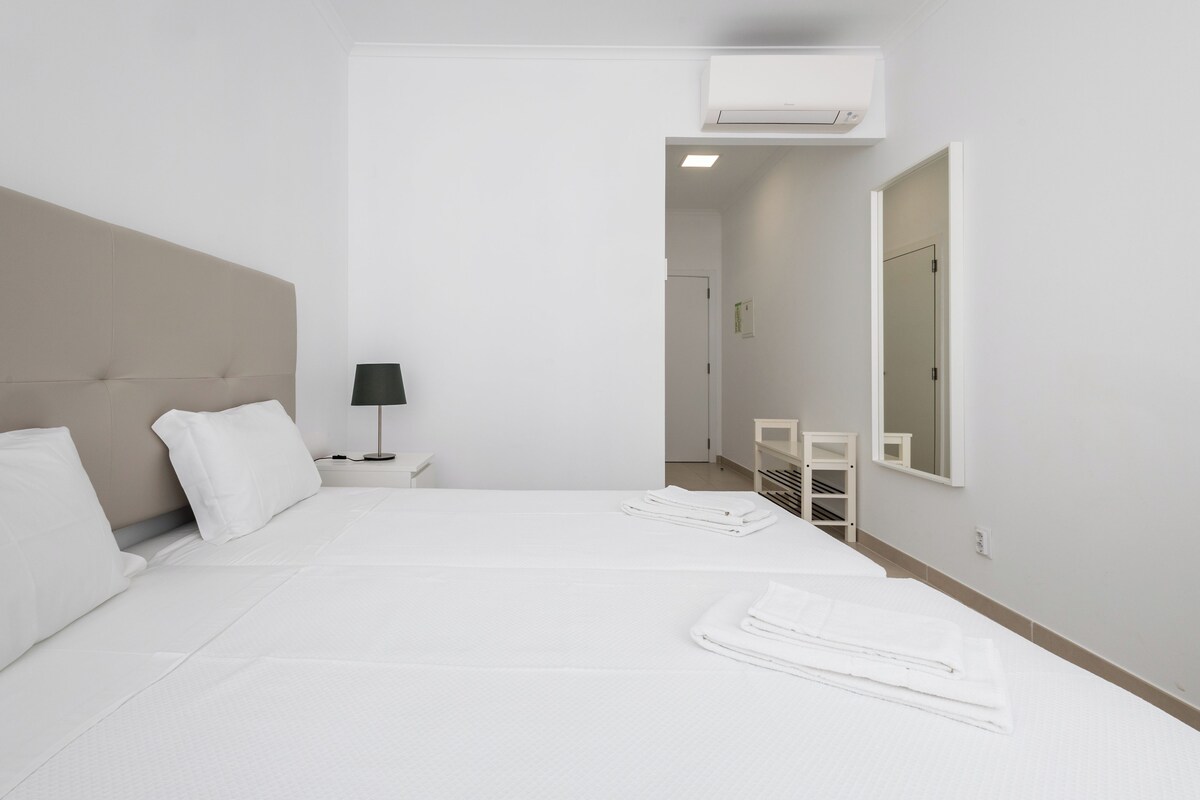 Habitación de hotel con una cama grande, una mesita de noche con lámpara, un espejo y un pequeño rincón de descanso con una silla y una estantería.