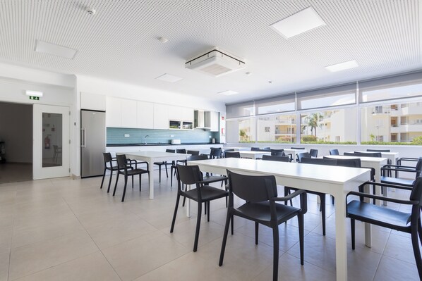 A spacious dining area with white tables and black chairs, large windows offering a view of the outside, and a modern kitchen area with stainless steel appliances. - Dunas Hostel & Guesthouse (Portimão)