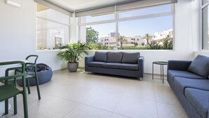 Un salon moderne avec un canapé gris, une petite table ronde et un fauteuil vert. Une grande fenêtre offre une vue sur les bâtiments et les arbres.