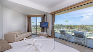 A modern hotel room with a large bed, a sofa, a TV, and a balcony with chairs and a view of the city.
