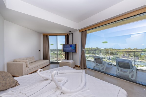 Habitación de hotel moderna con una cama grande, un sofá, un televisor y un balcón con sillas y vistas a la ciudad.