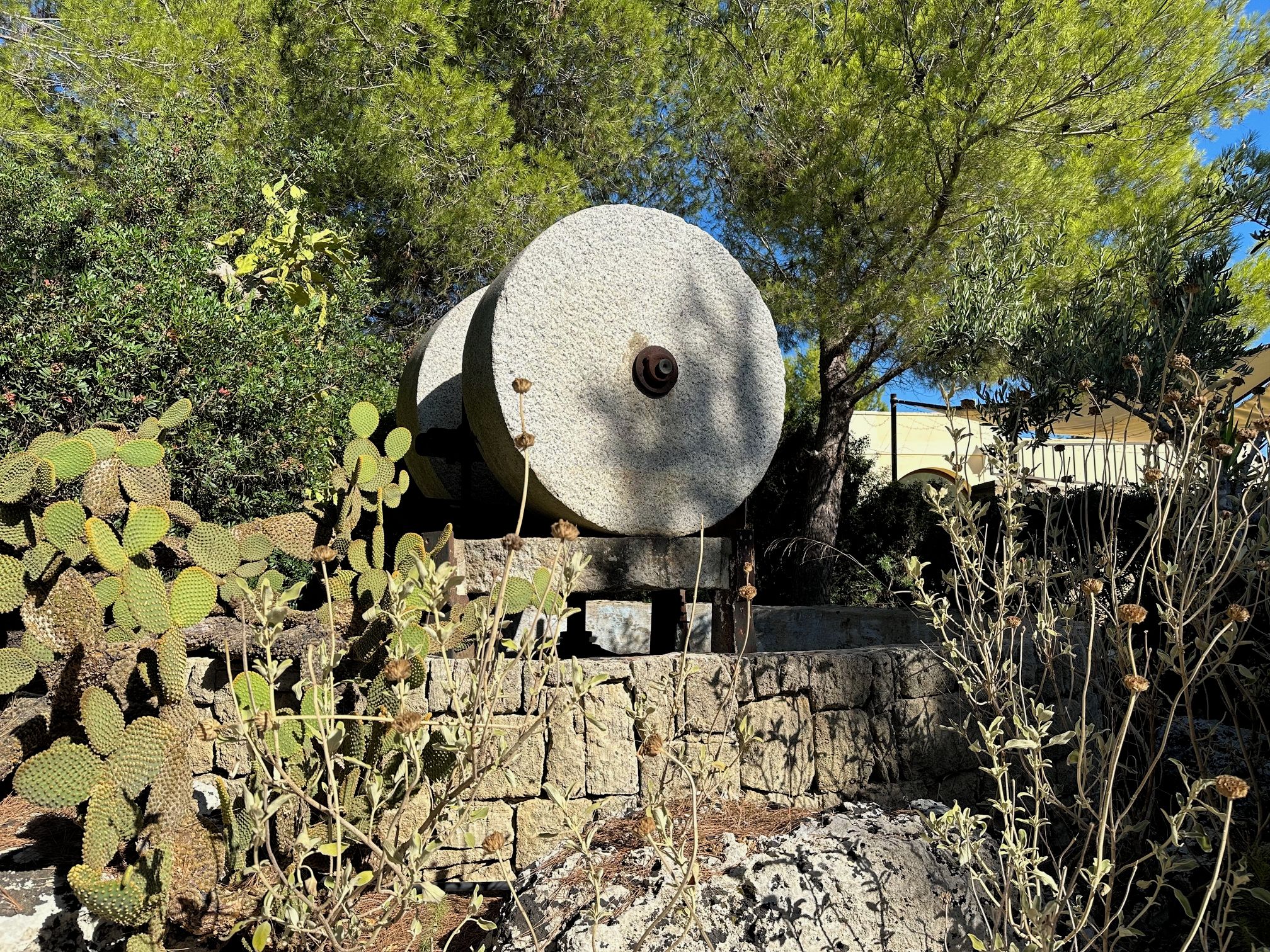 Une grande structure cylindrique en pierre, surmontée d’un boulon rouillé, entourée de différentes plantes et d’un mur de pierre.