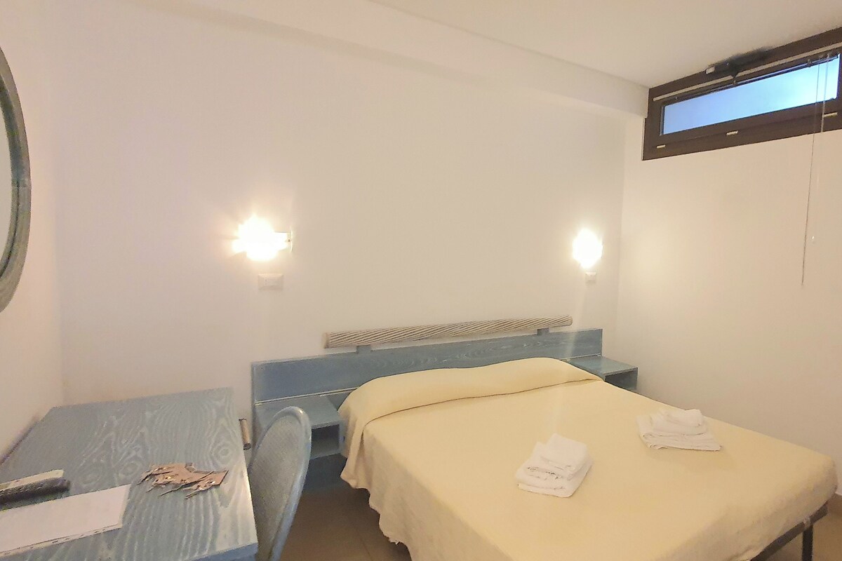 A small, simple room with a bed, a desk, and a mirror. There are two wall-mounted lights and a window with a blind.