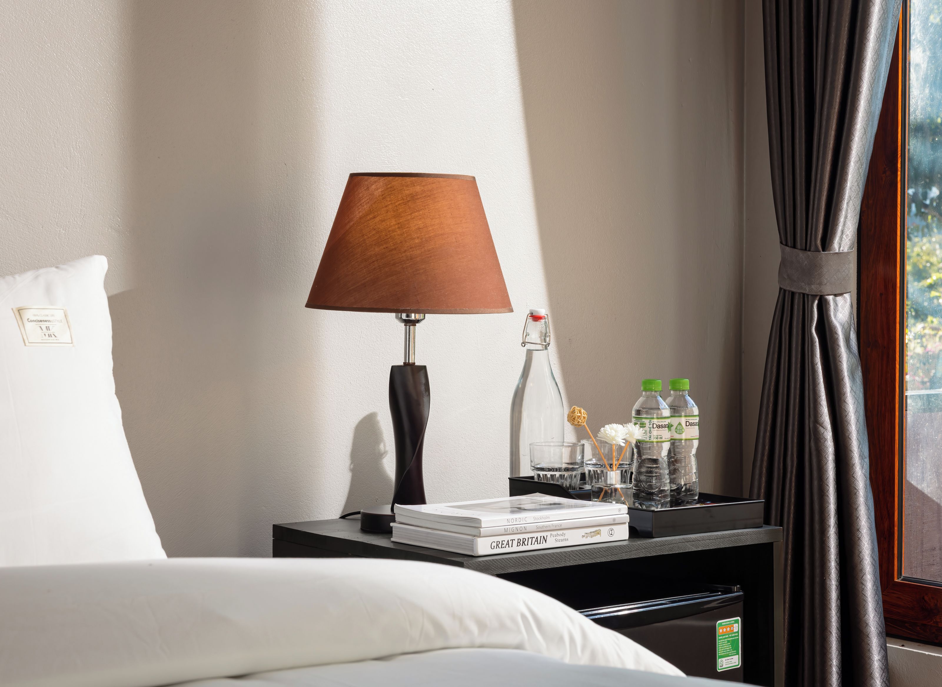 Una habitación de hotel impecable con una cama, una mesita de noche con lámpara, una botella y dos botellas de agua, además de una pila de libros.