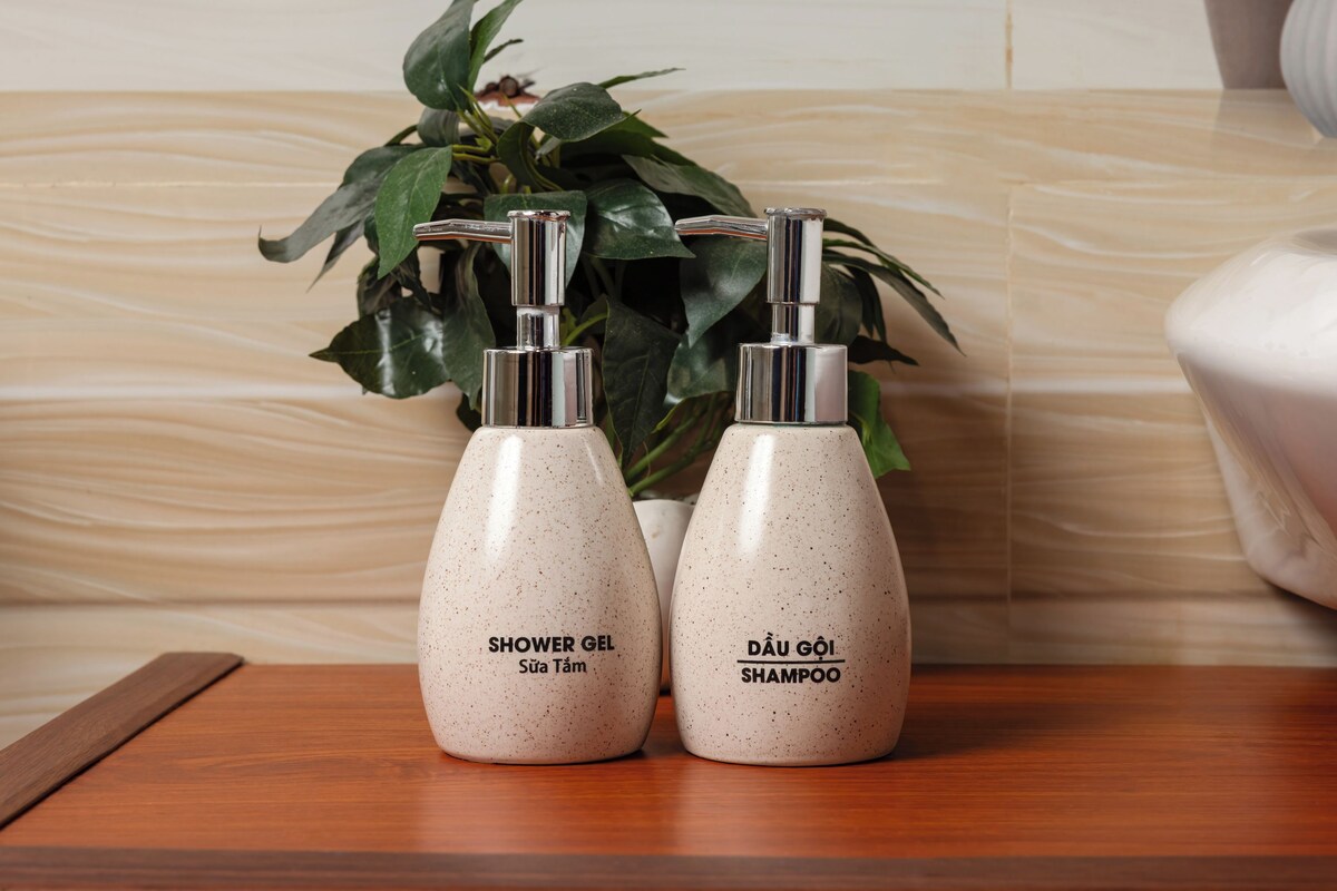 Dos botellas de artículos de aseo personal etiquetadas 'Shower Gel' y 'Champú' sobre una superficie de madera, con una planta de fondo.