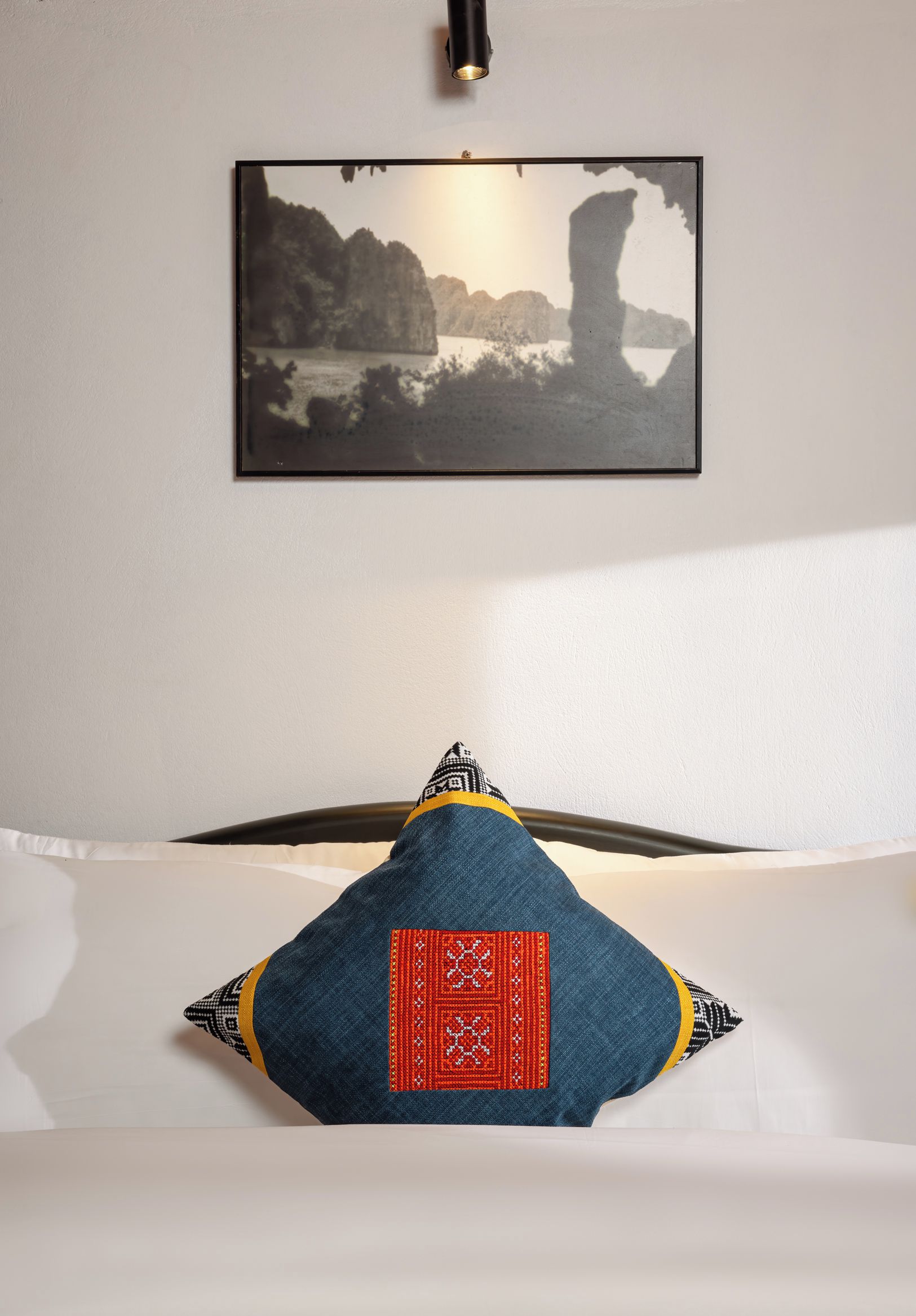 A bed with a pillow featuring a red and black pattern, a framed picture of a coastal landscape on the wall, and a ceiling light fixture.