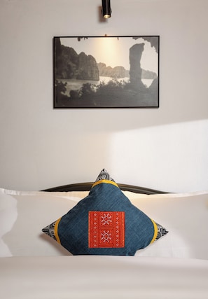 Un lit avec un oreiller orné d’un motif rouge et noir, un tableau encadré représentant un paysage côtier accroché au mur et un luminaire fixé au plafond.
