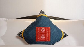 Un lit avec un oreiller orné d’un motif rouge et noir, un tableau encadré représentant un paysage côtier accroché au mur, et un luminaire placé au-dessus du lit.