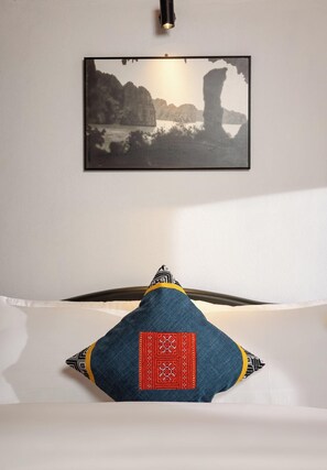 A bed with a pillow featuring a red and black pattern, a framed picture of a coastal landscape on the wall, and a light fixture above the bed.
