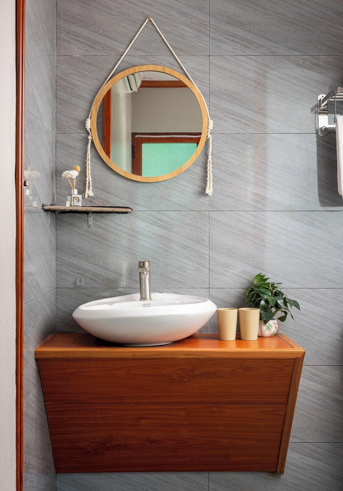 A bathroom with a wooden vanity, a white sink, a round mirror with a rope handle, and a potted plant.