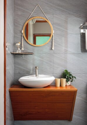 Un bagno con un mobile contenitore in legno, un lavandino bianco, uno specchio rotondo con maniglia in corda e una pianta in vaso.