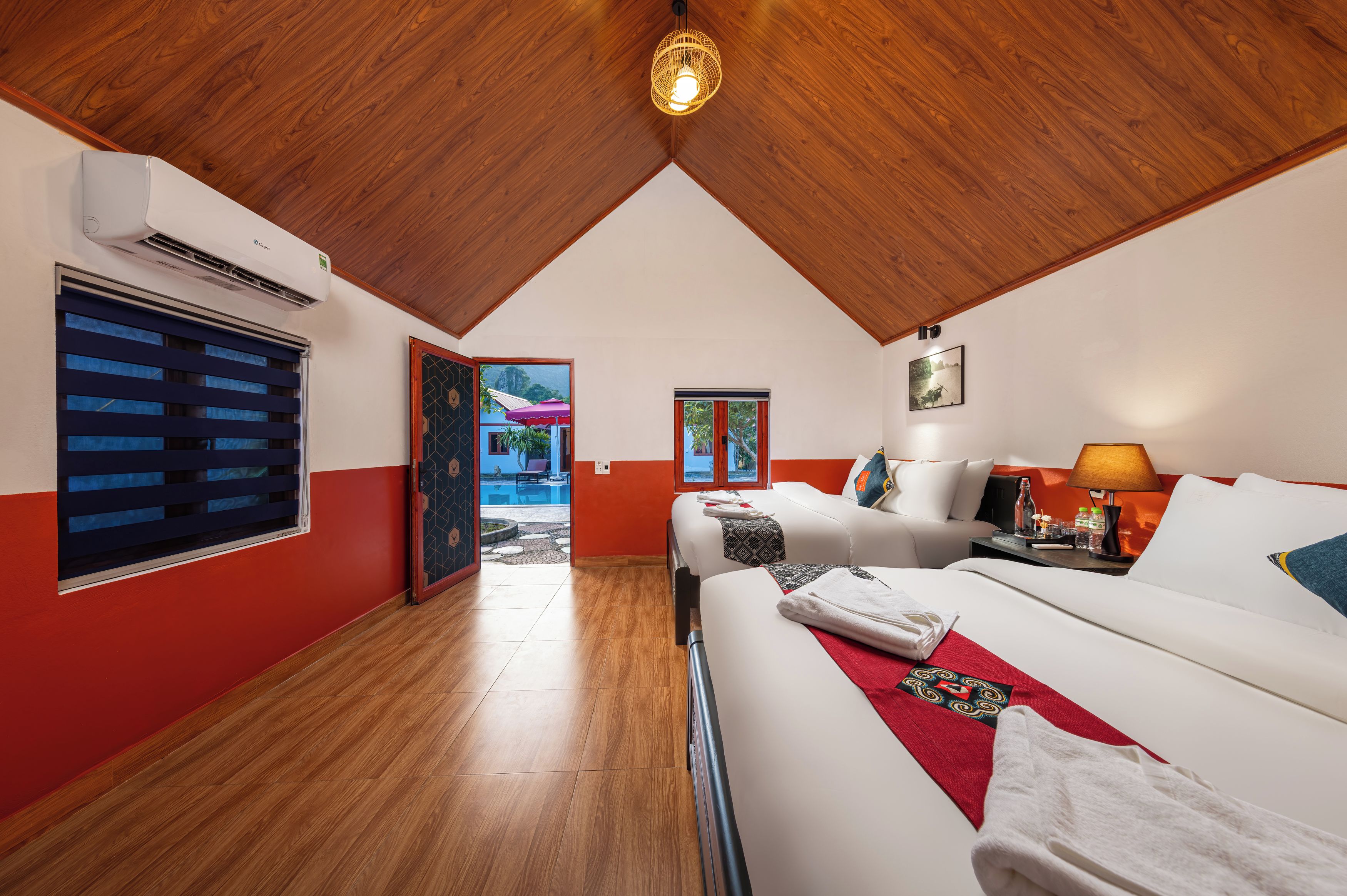 A hotel room with a wooden ceiling, a bed with white linens, a red accent wall, and a window with blinds.