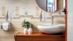 A bathroom with a wooden vanity, a round mirror, and a white sink. There are towels hanging on a rack, a vase with flowers, and a small potted plant on the vanity. - Cat Ba Serena Homestay And Swimming Pool (Cat Hai)