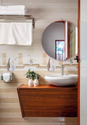 A bathroom with a wooden vanity, a round mirror, and a white sink. There are towels hanging on a rack, a vase with flowers, and a small potted plant on the vanity. - Cat Ba Serena Homestay And Swimming Pool (Cat Hai)