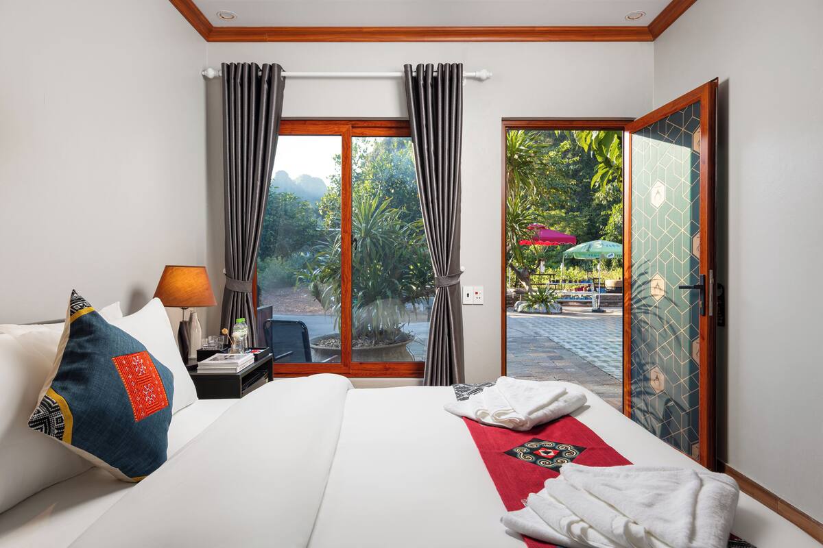 Une chambre d’hôtel avec un lit, une table de chevet, une lampe et une vue sur un espace extérieur agrémenté de plantes et de parasols.