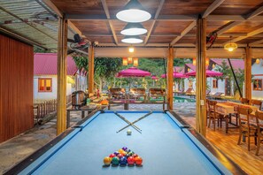Een pooltafel met biljartballen en stokken, omringd door houten meubels en tafels.
