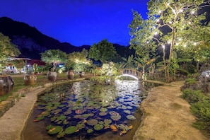 Ein Garten bei Nacht mit Teich, beleuchteten Pflanzen und einer Holzbrücke.