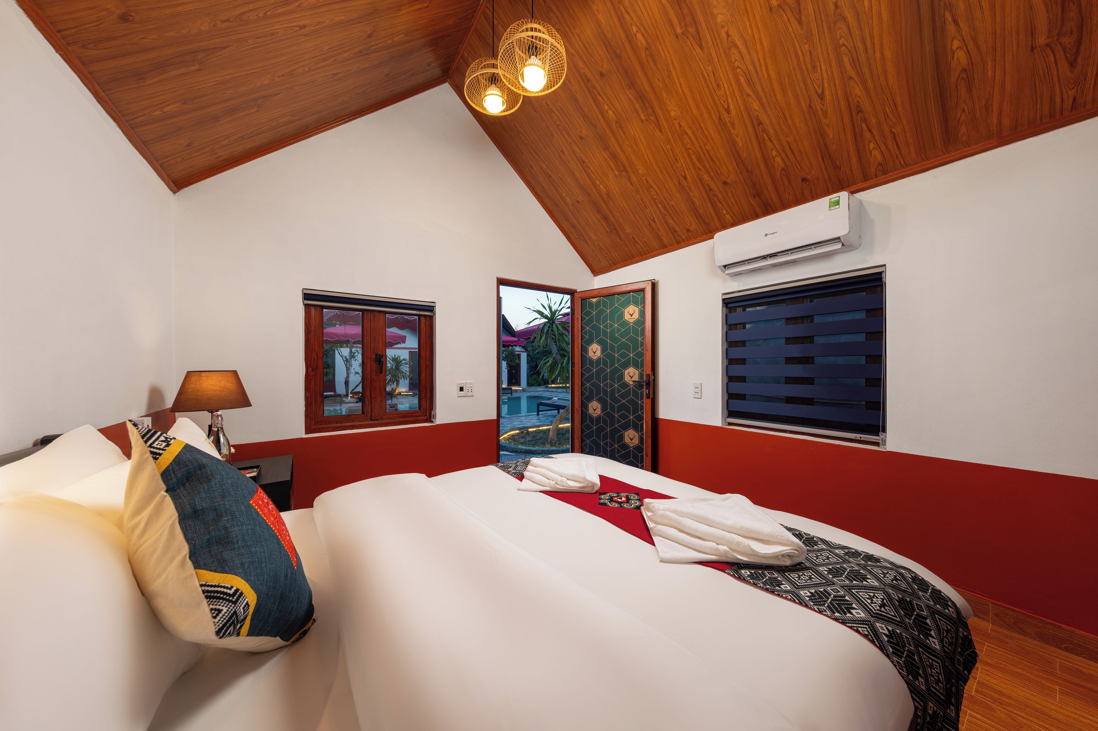 A hotel room with a wooden ceiling, a bed with white bedding, a red wall, and a window with blinds.